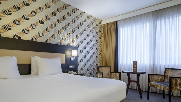 A hotel room with a large bed, a small table, two chairs, a nightstand, and a patterned wallpaper. - Mercure Antwerp City South (Antwerp)