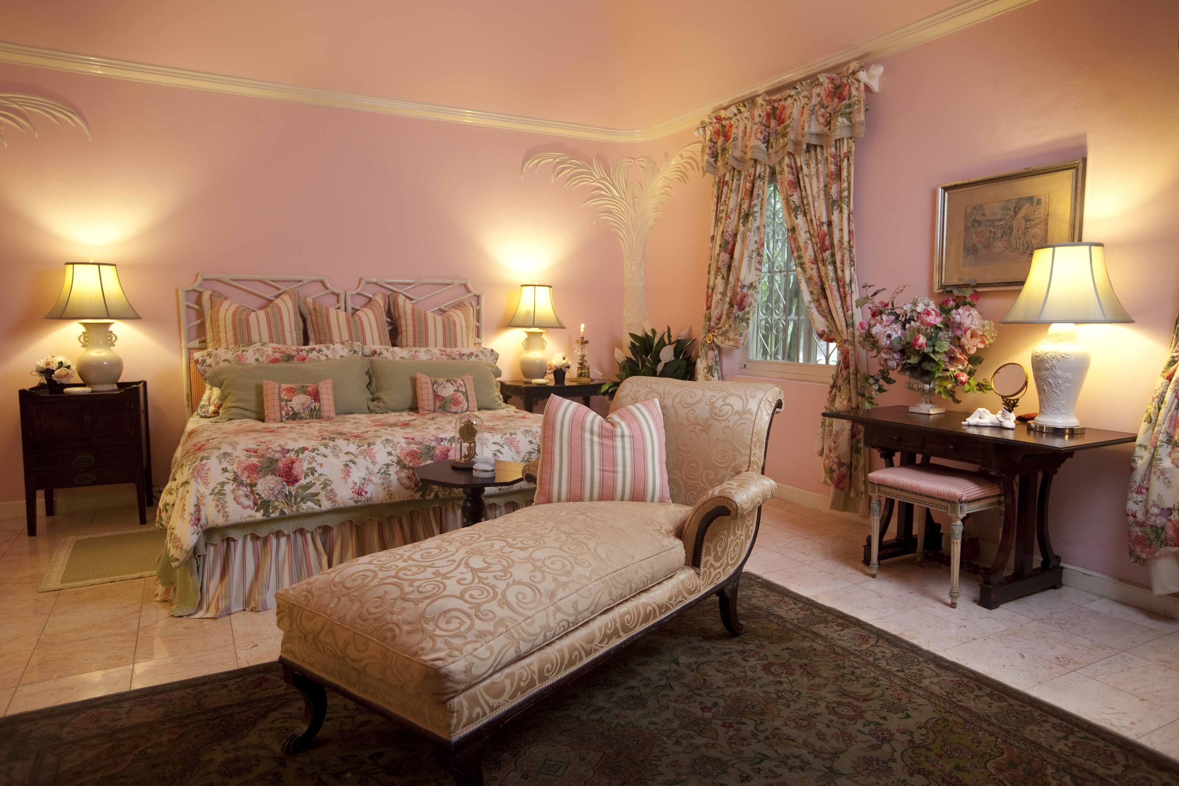 A bedroom with a large bed, a chaise lounge, a desk, and a chair. There are lamps on both sides of the bed and a window with curtains.