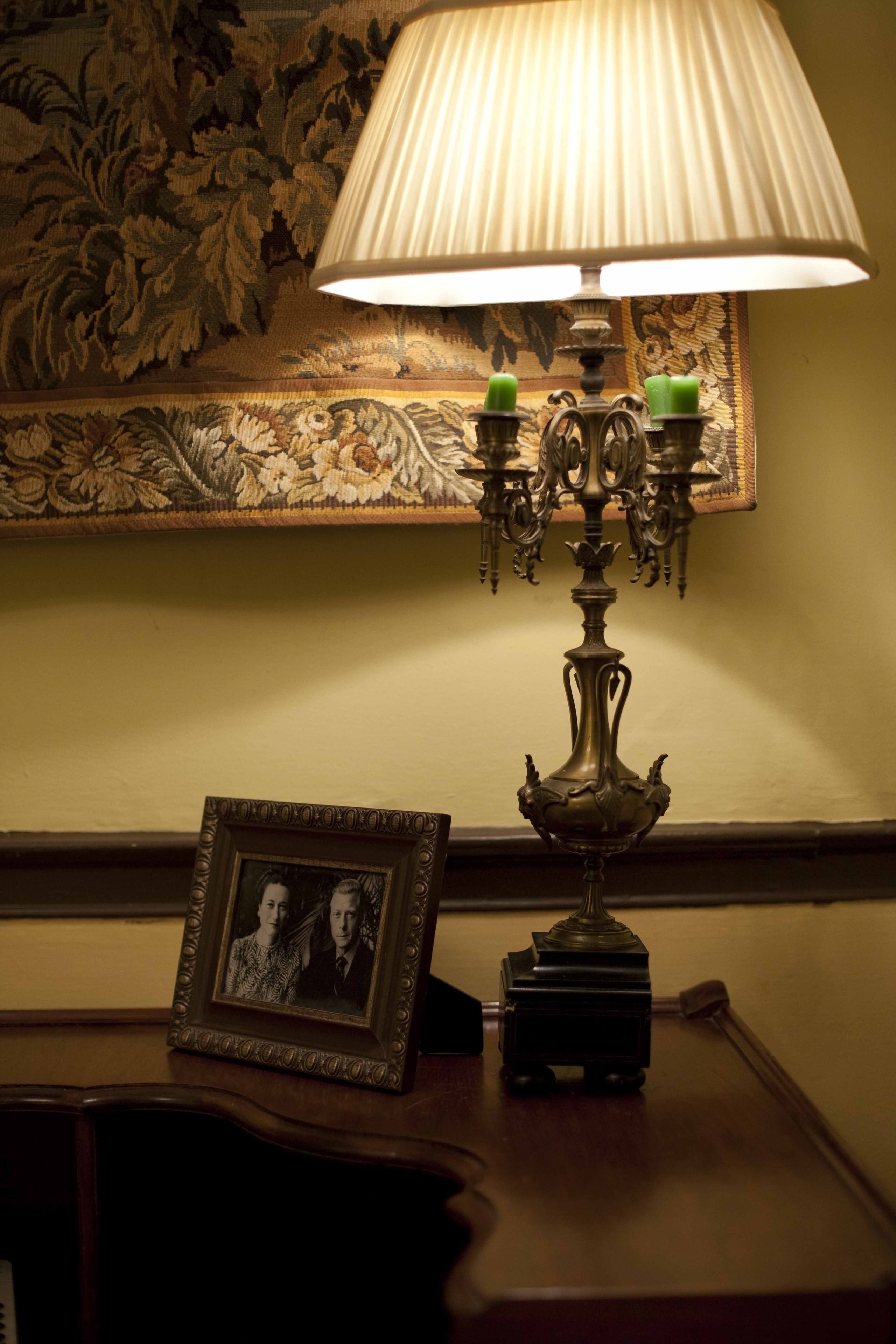 A table with a lamp, a framed photograph, and a floral tapestry on the wall.