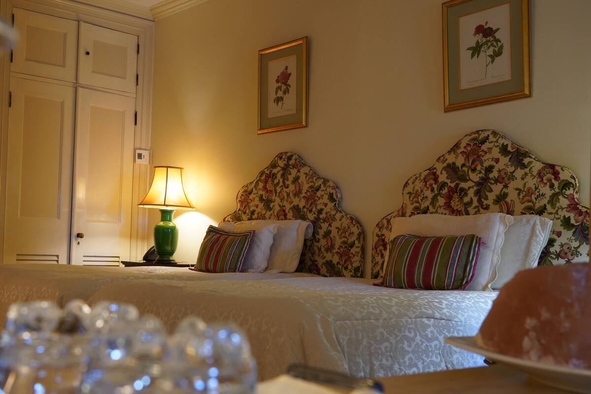 A hotel room with two beds, floral headboards, a lamp, and framed botanical prints on the wall.