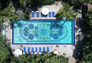 Vista aérea de una piscina rectangular con un diseño de mosaicos decorativos, rodeada de palmeras y sillas de descanso azules.