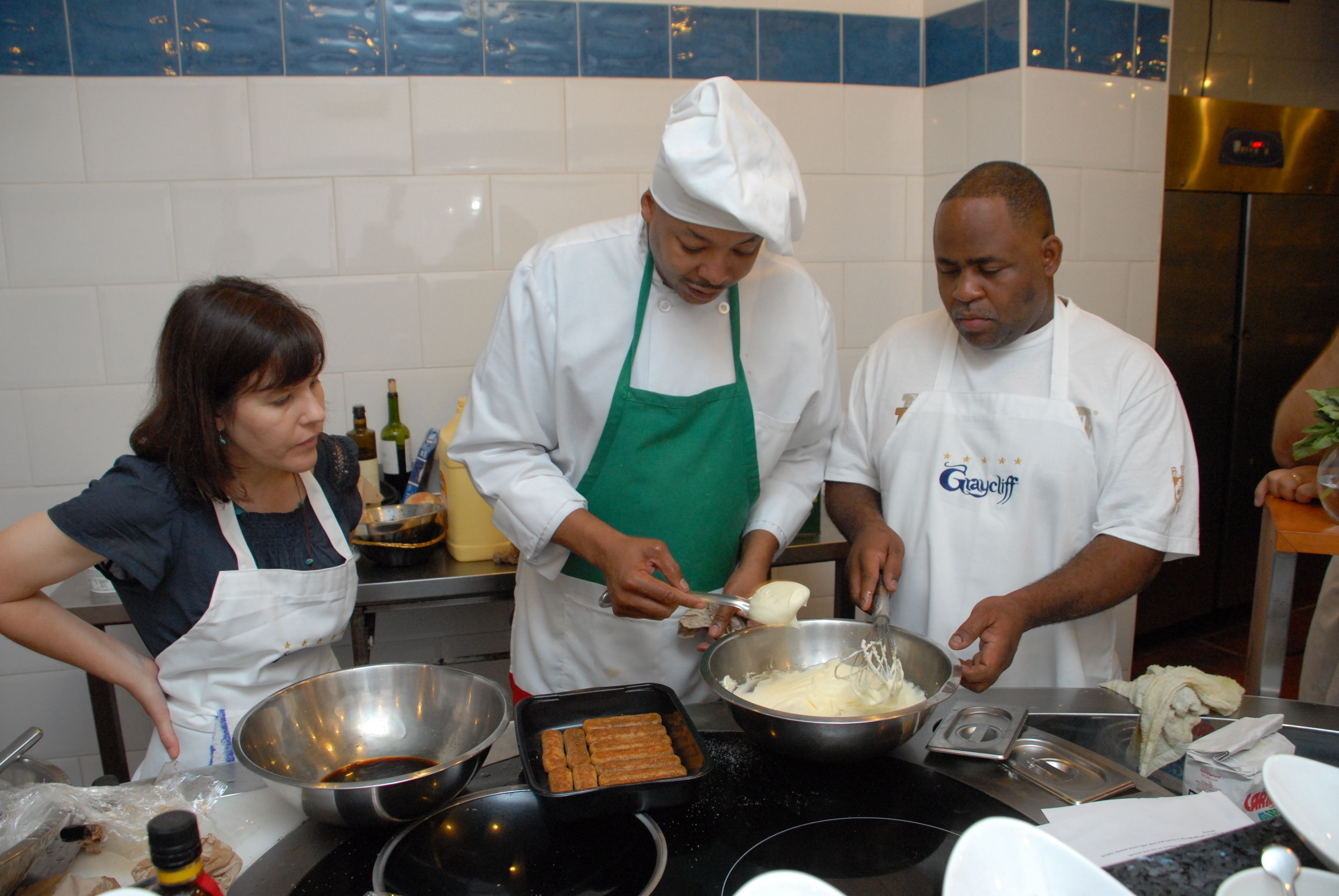A kitchen with three people preparing food, various cooking utensils, and ingredients.
