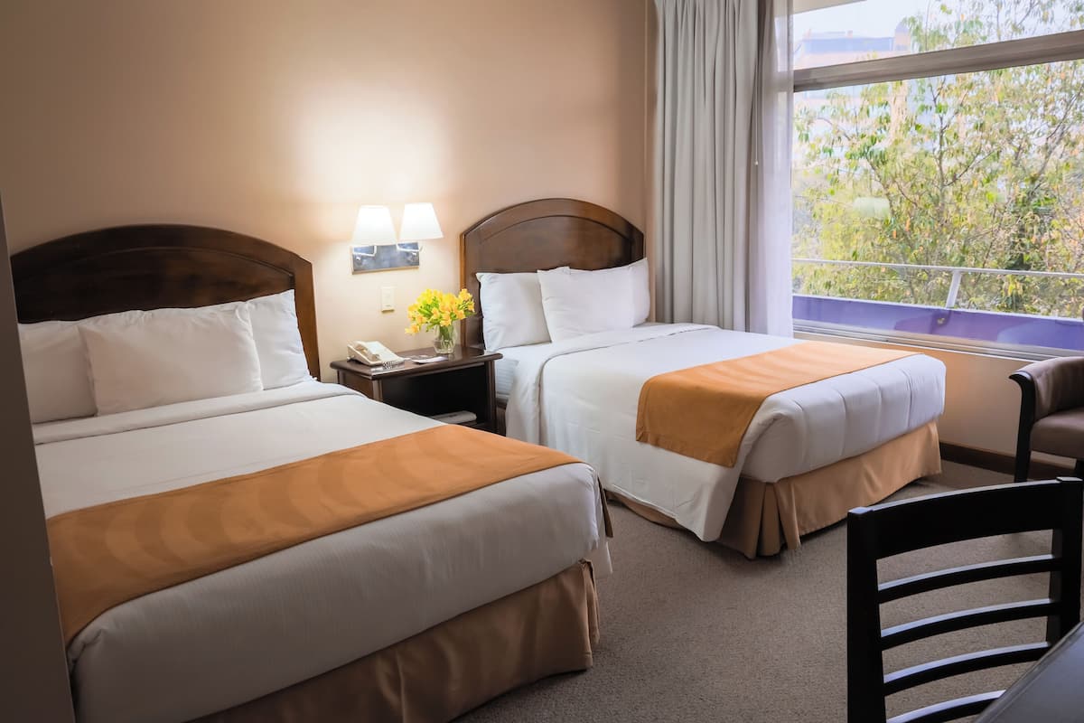 A hotel room with two beds, a nightstand with a lamp, a chair, and a window with a view of trees.