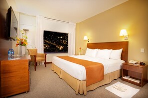 A hotel room with a large bed, a desk, a chair, a nightstand, a lamp, a vase with flowers, and a window with a city view at night.