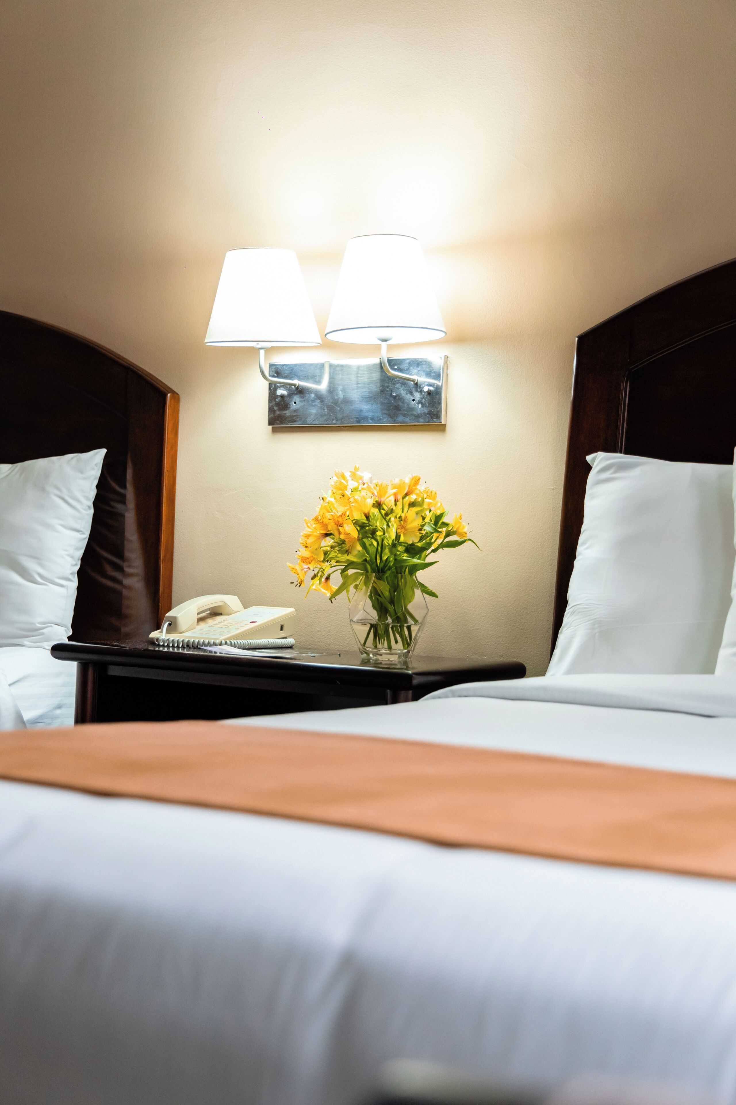 Una habitación de hotel con una cama, una mesita de noche con una lámpara, un teléfono y un florero con flores.