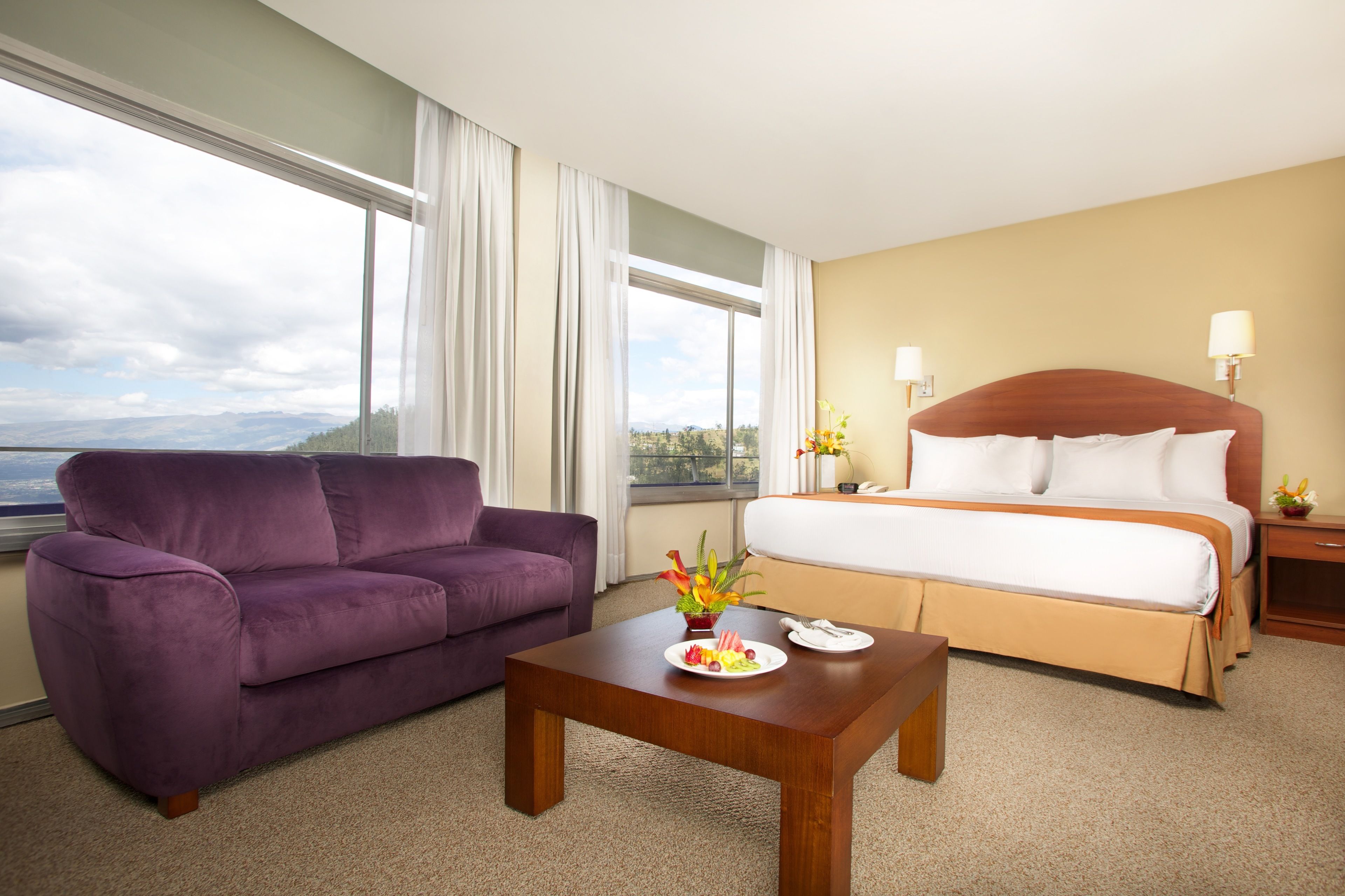 A hotel room with a bed, a purple sofa, a wooden coffee table with a plate of fruit, and a view of the mountains through large windows.