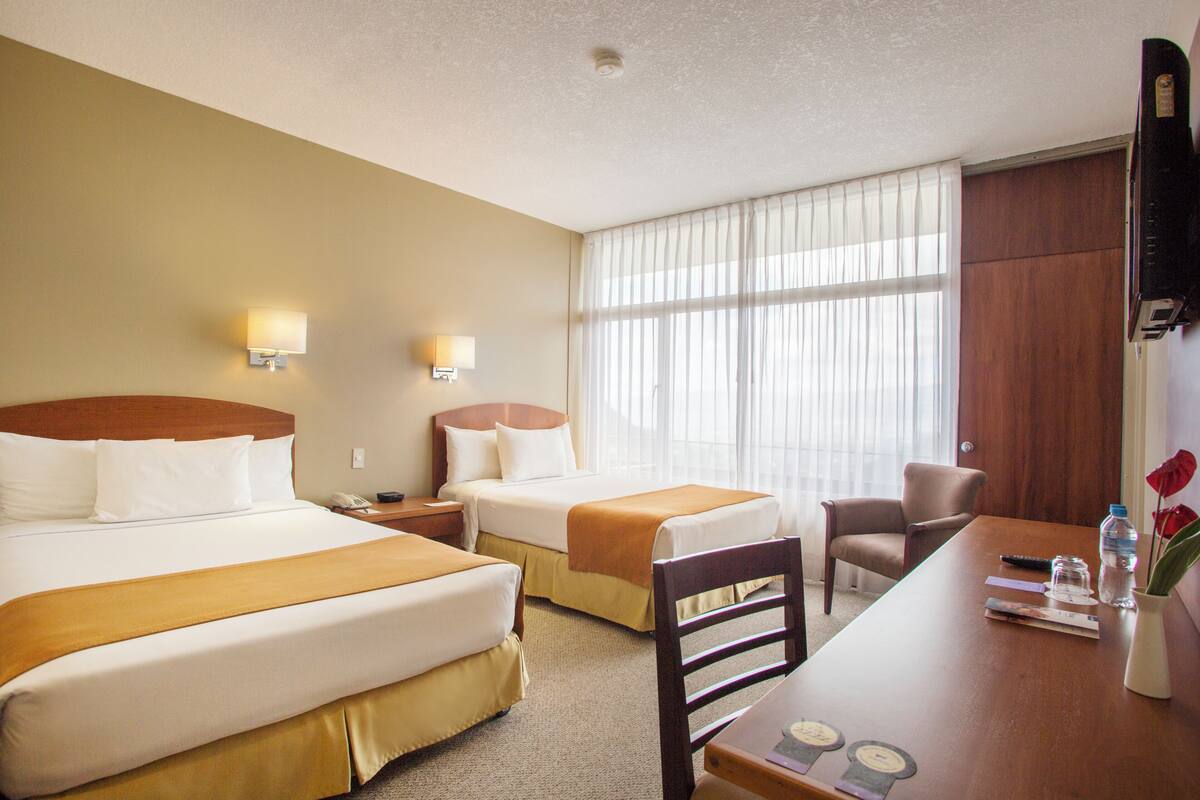 A hotel room with two beds, a desk, a chair, a TV, and a window with curtains.