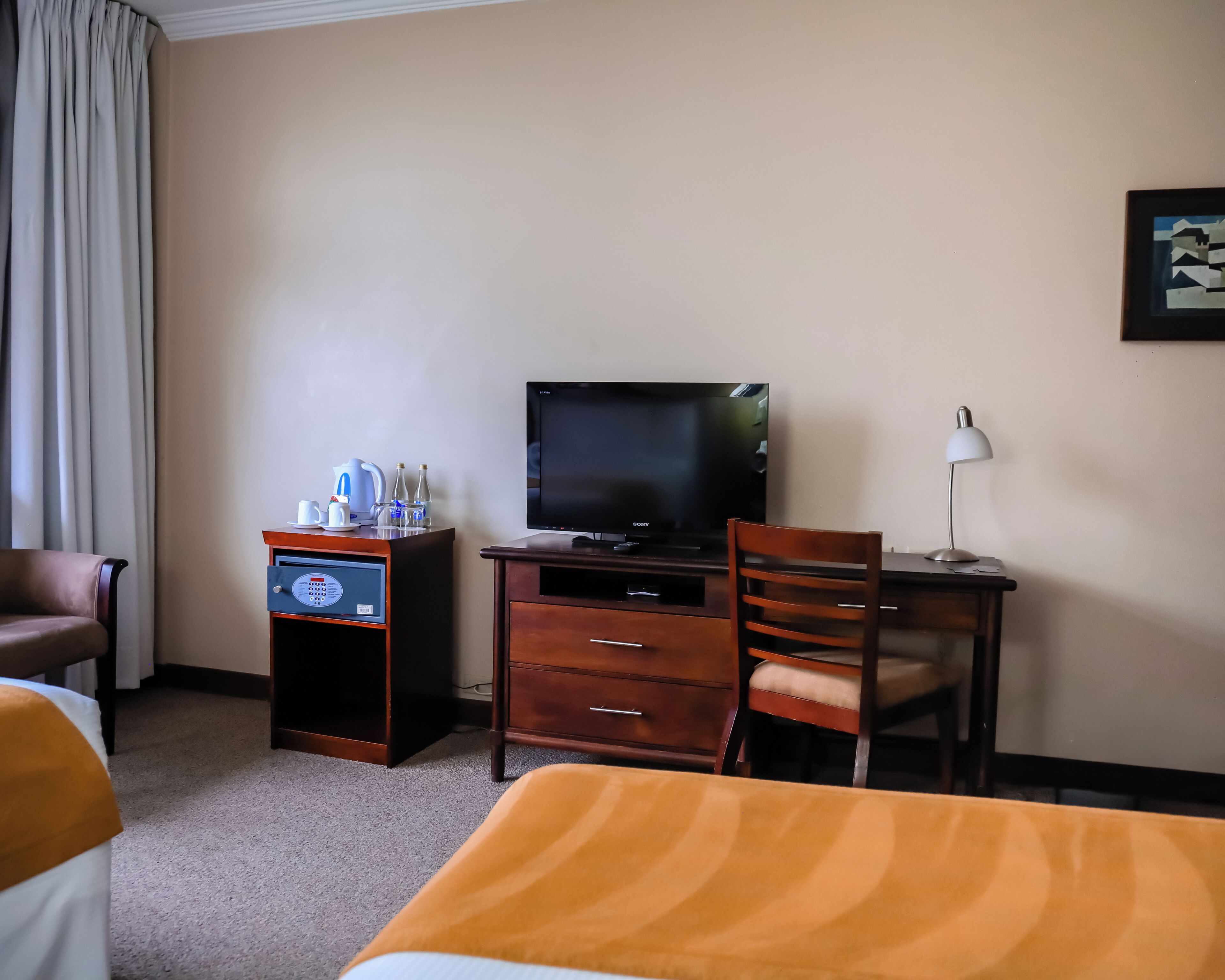 A hotel room with a bed, a desk with a television, a chair, a desk lamp, and a minibar.