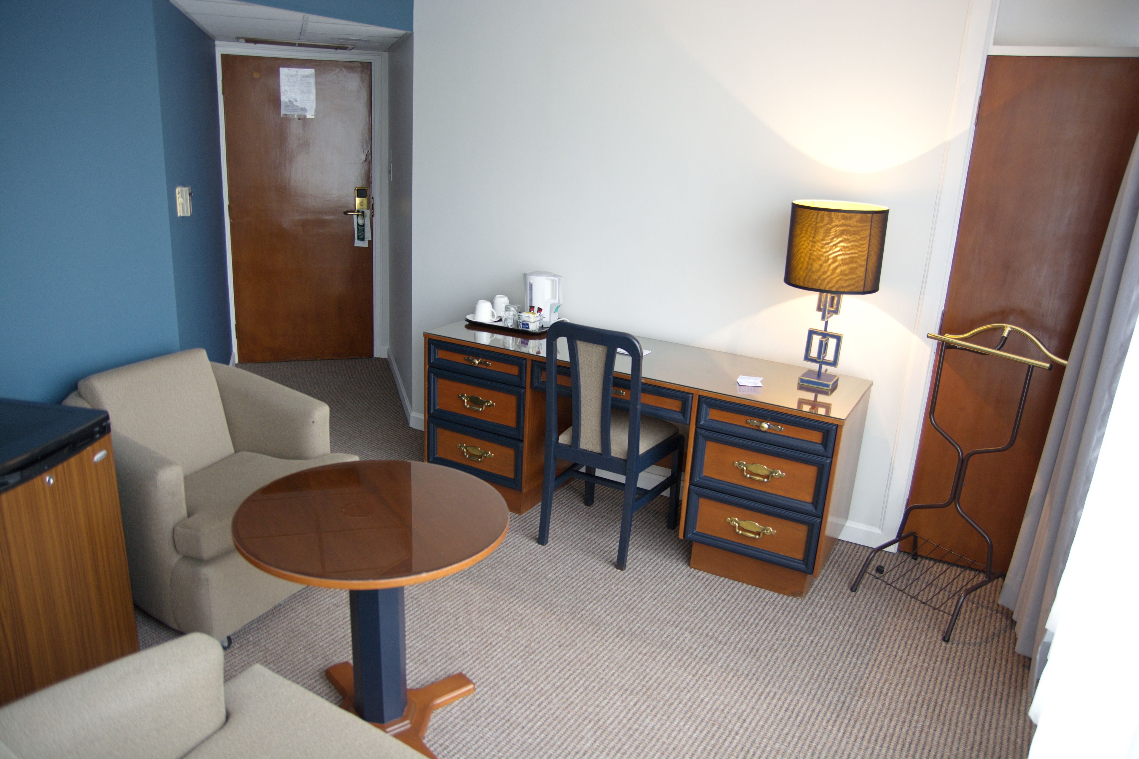 Habitación de hotel compacta con escritorio, silla y una mesita. Hay una lámpara en el escritorio y un perchero cerca de la puerta.