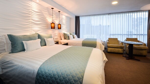Habitación de hotel con dos camas, un sofá, una mesita y vistas a la ciudad a través de amplios ventanales.