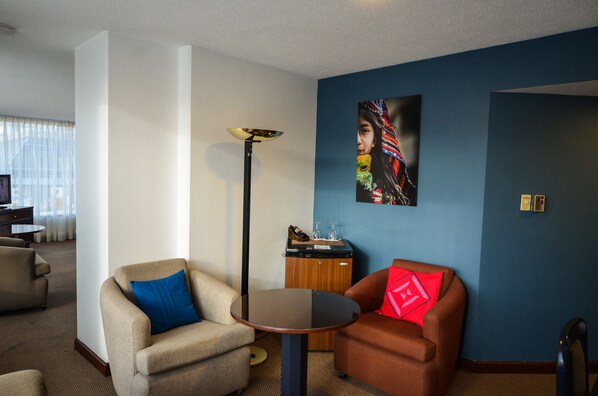 Habitación de hotel con dos sillones, una mesita redonda, una lámpara de pie y un televisor instalado en la pared.