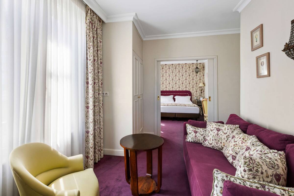 A hotel room with a yellow armchair, a purple sofa, a wooden side table, and a bed with floral-patterned bedding.