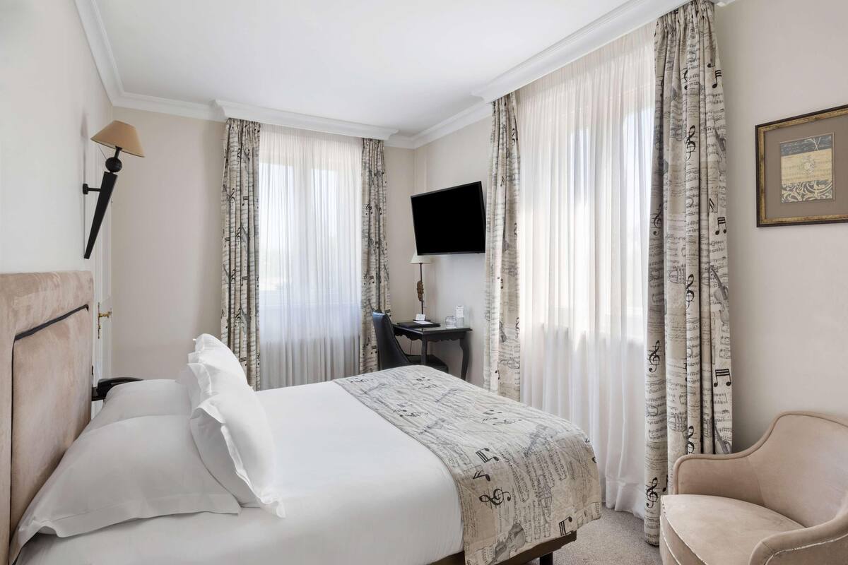 A hotel room with a bed, a desk with a flat-screen TV, a chair, and curtains with a musical note pattern.