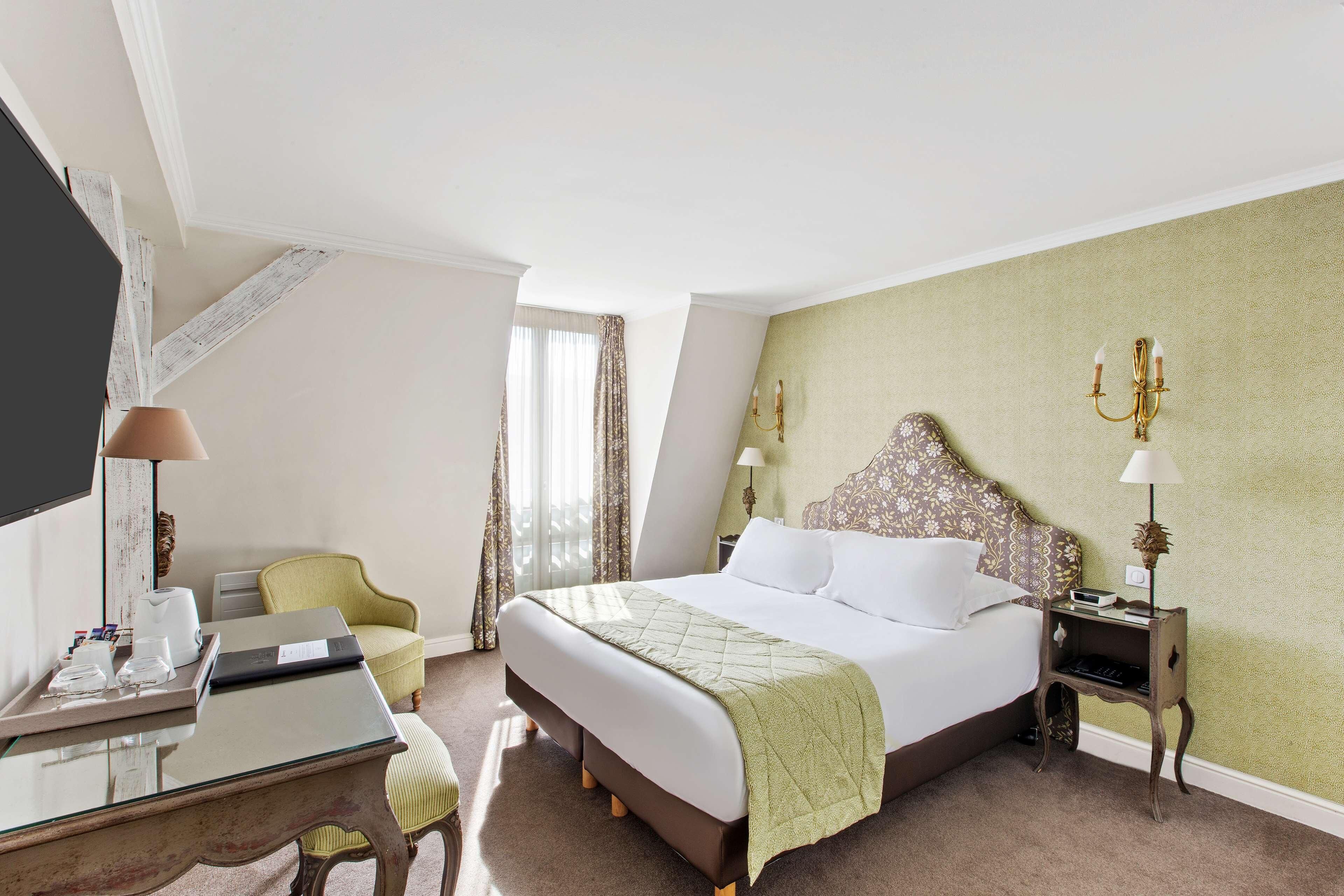 Quarto de hotel com cama, escrivaninha, cadeira, mesa de cabeceira e luminária.