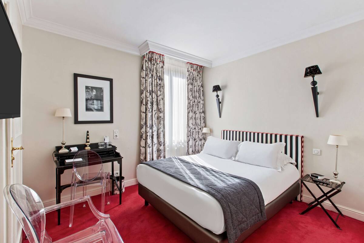 A hotel room with a large bed, a desk with a chair, a small table, and two wall-mounted lamps.