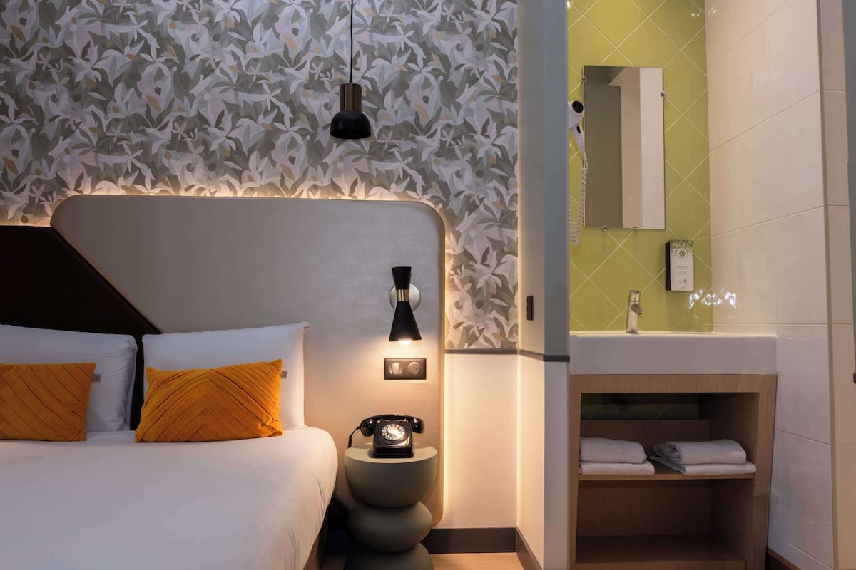 Een hotelkamer met een bed, een nachtkastje met een draaischijftelefoon, een wandlamp, een spiegel en een kaptafel met een handdoekhouder.