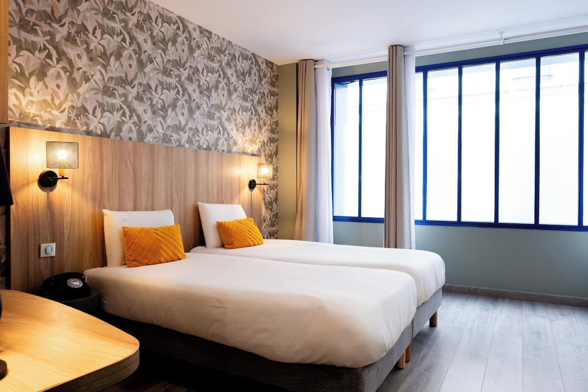 Hotelkamer met twee bedden, een houten hoofdbord, een behang met patroon en een groot raam met gordijnen.
