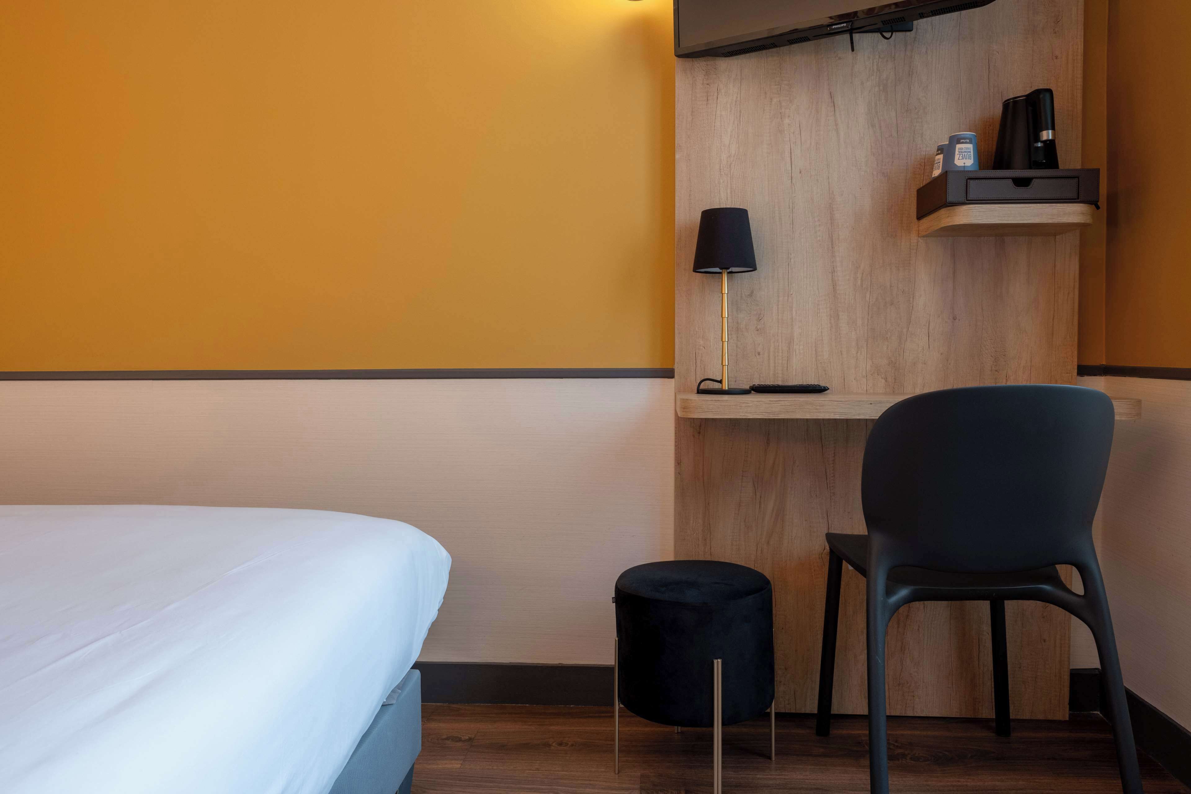 A hotel room with a bed, a small black chair, a wooden shelf with a lamp, and a trash bin.
