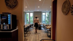 A modern indoor space with a coffee machine, tables, chairs, and large windows offering a view of greenery. - Hotel Kyriad Paris 10 Canal Saint Martin (Paris)
