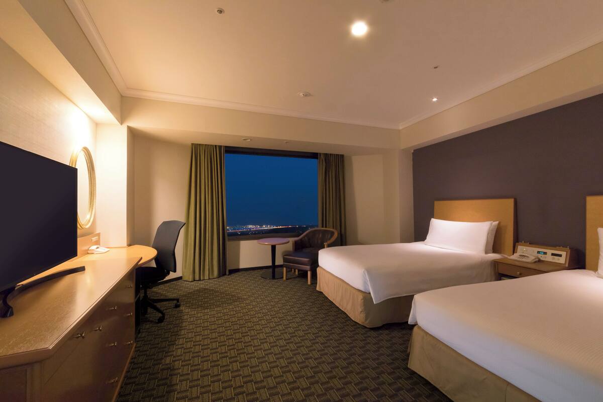 Habitación de hotel con dos camas, un escritorio con computadora, una silla, una mesita y vista a la ciudad de noche.