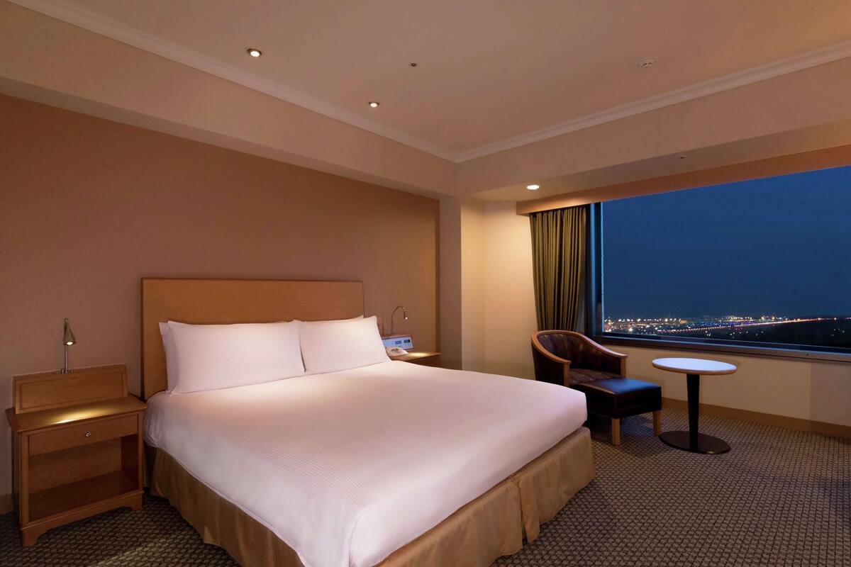 Habitación de hotel con una cama grande, una mesita de noche, una silla, una pequeña mesa y una ventana con vista a la ciudad.