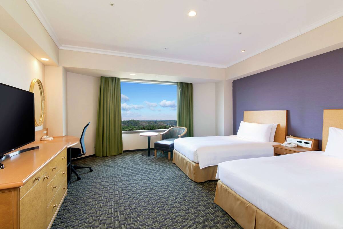Habitación de hotel con dos camas, un escritorio con televisor, una silla, una mesita y una ventana con vistas al paisaje.