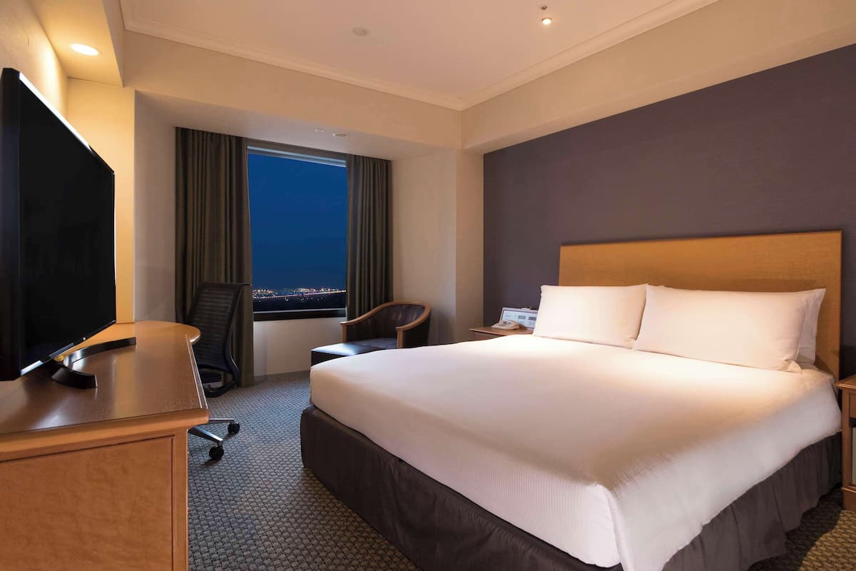 Habitación de hotel con una cama grande, un escritorio con computadora, una silla, un televisor y una ventana con vista a la ciudad de noche.