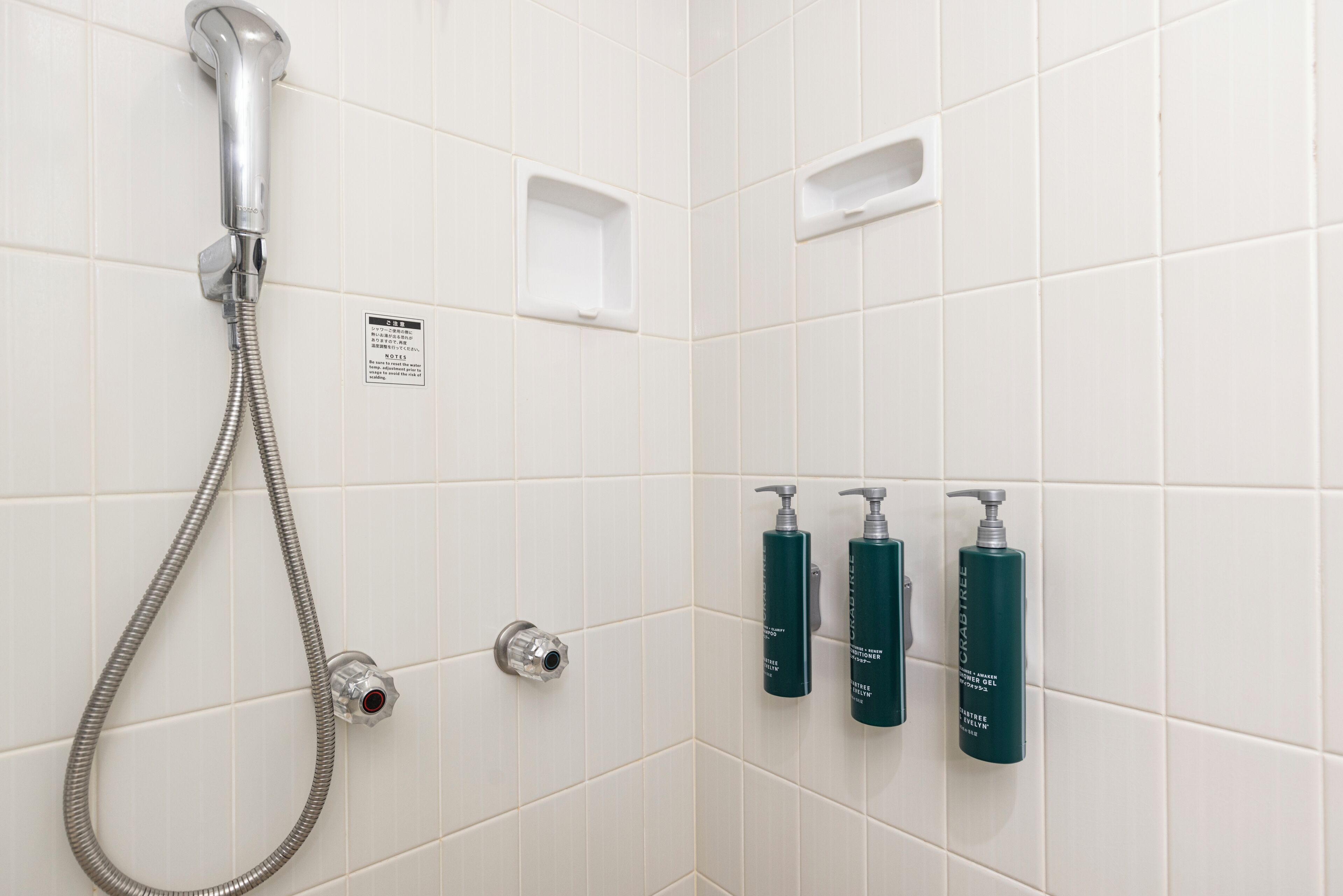 A bathroom with white tiles, a shower with a handheld hose, and three green bottles of shampoo and conditioner on the wall.