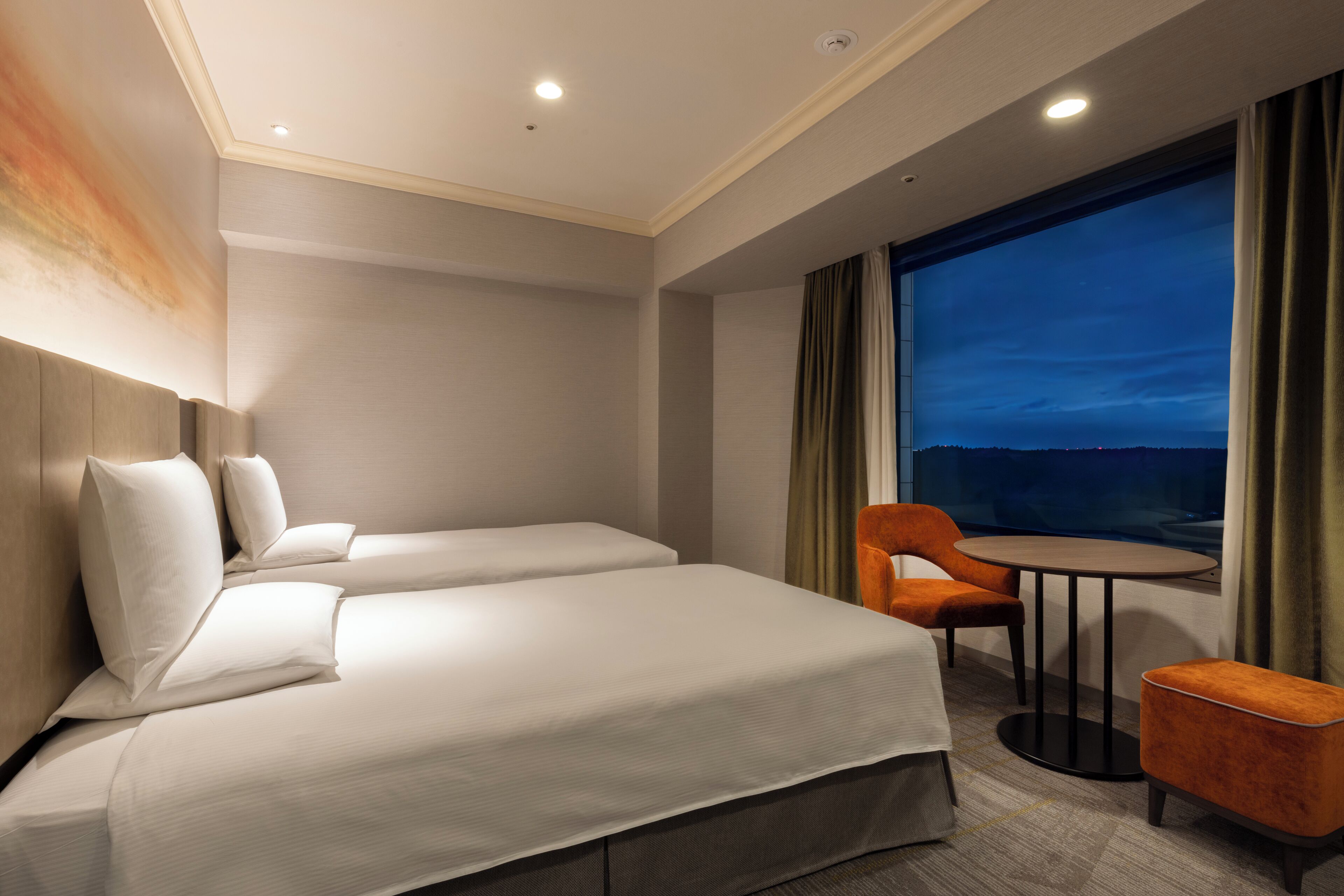 Habitación de hotel con dos camas, una mesa redonda y vistas a la ciudad al atardecer.