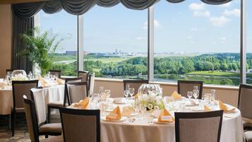 A dining area with tables set for a meal, large windows with a view of a landscape, and a potted plant.