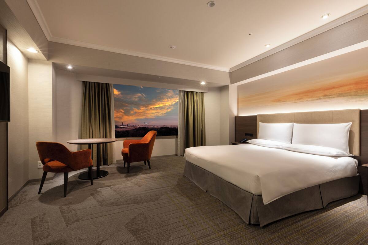 Una habitación de hotel moderna con una cama grande, dos sillas, una mesita y vistas panorámicas por la ventana.