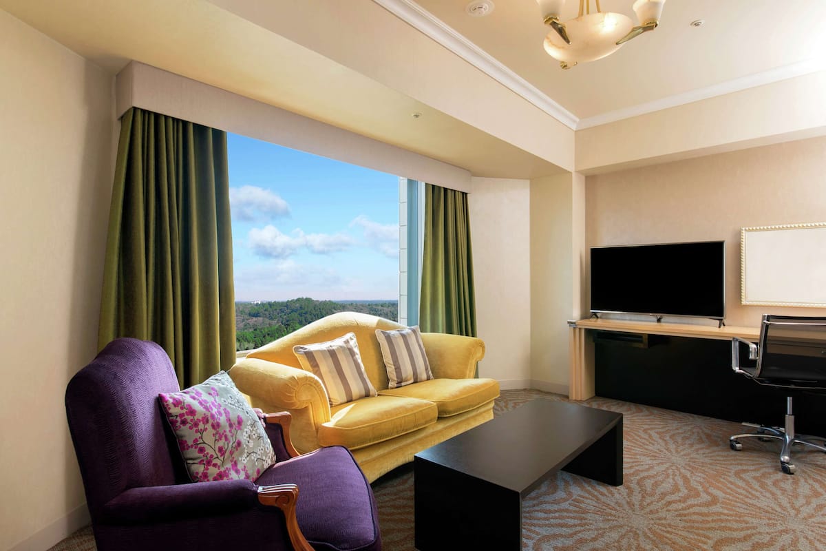 Habitación de hotel con un sofá amarillo, un sillón púrpura, una mesa de centro negra, un televisor y vistas al exterior.