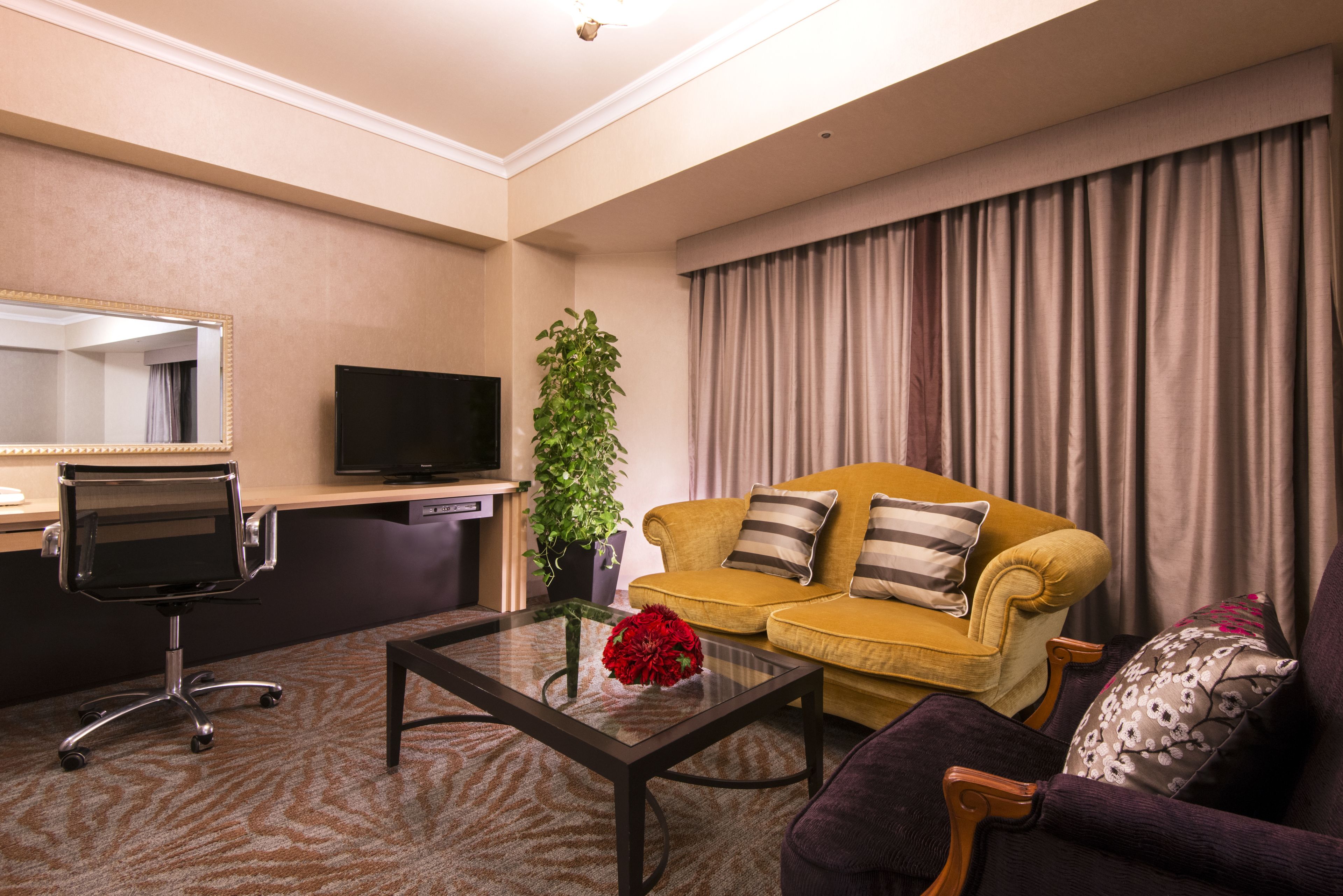A hotel room with a sofa, armchair, TV, desk, and a potted plant.
