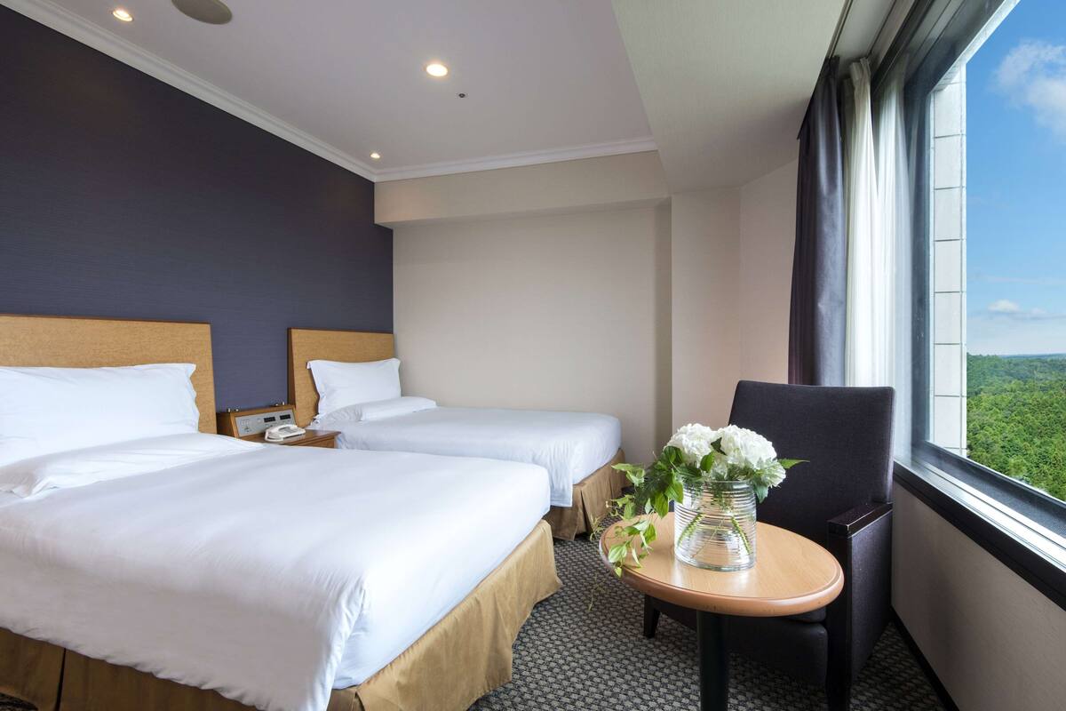 Habitación de hotel con dos camas, una mesita con un florero y una ventana con vistas a zonas verdes.