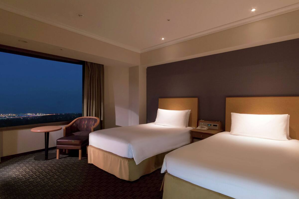 Habitación de hotel con dos camas, una silla, una mesita y vista a la ciudad de noche.