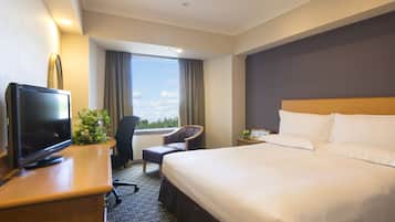 Habitación de hotel con una cama grande, un escritorio con televisor, una silla y vistas a la ciudad desde la ventana.