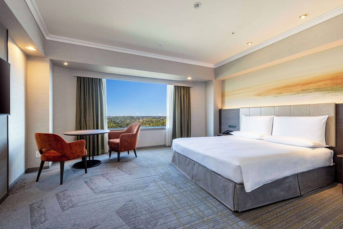 Habitación de hotel con una cama grande, un escritorio, dos sillas, vistas al exterior y un cuadro en la pared.