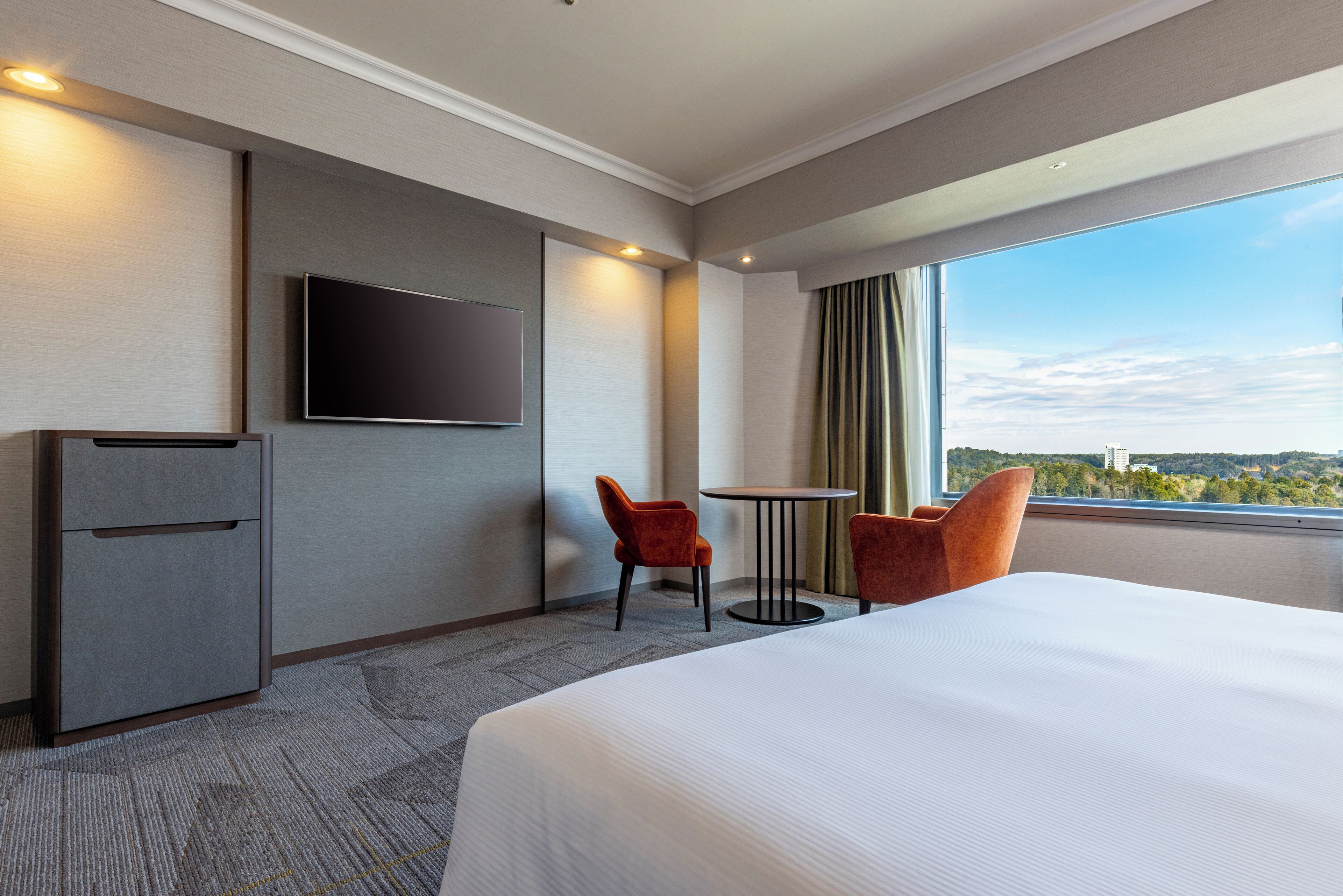 Habitación de hotel con una cama grande, televisión montada en la pared, una mesita pequeña, dos sillas y vistas a la ciudad por la ventana.