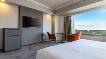 Habitación de hotel con una cama grande, televisión montada en la pared, una mesita pequeña, dos sillas y vistas a la ciudad por la ventana.