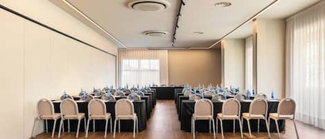 A conference room with rows of tables and chairs, equipped with water bottles and microphones, featuring a wooden floor and large windows.