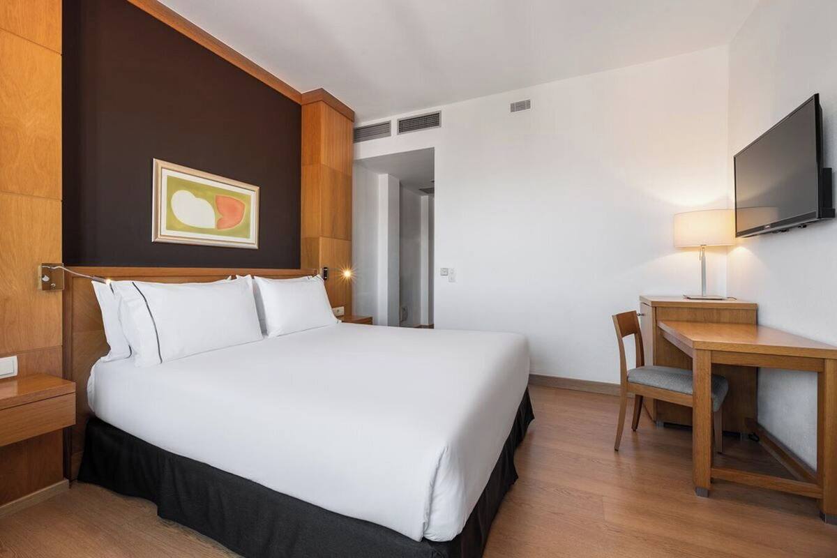 A hotel room with a bed, a desk, a chair, a TV, and a painting on the wall.