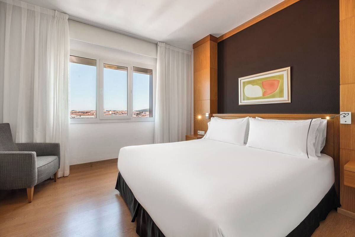 A hotel room with a large bed, a chair, a window with curtains, and a painting on the wall.