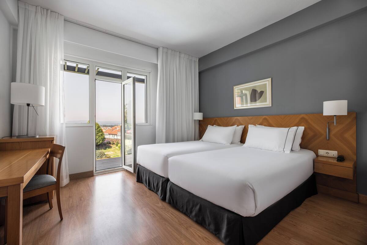 A hotel room with two beds, a desk, and a chair. There is a window with curtains, a lamp, and a framed picture on the wall.