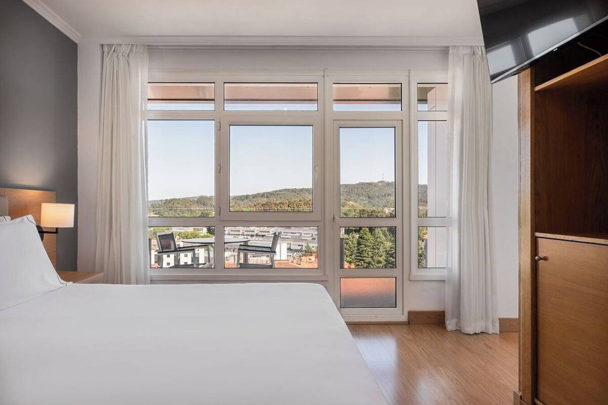 A hotel room with a large bed, a view of the city through floor-to-ceiling windows, and a wooden wardrobe.