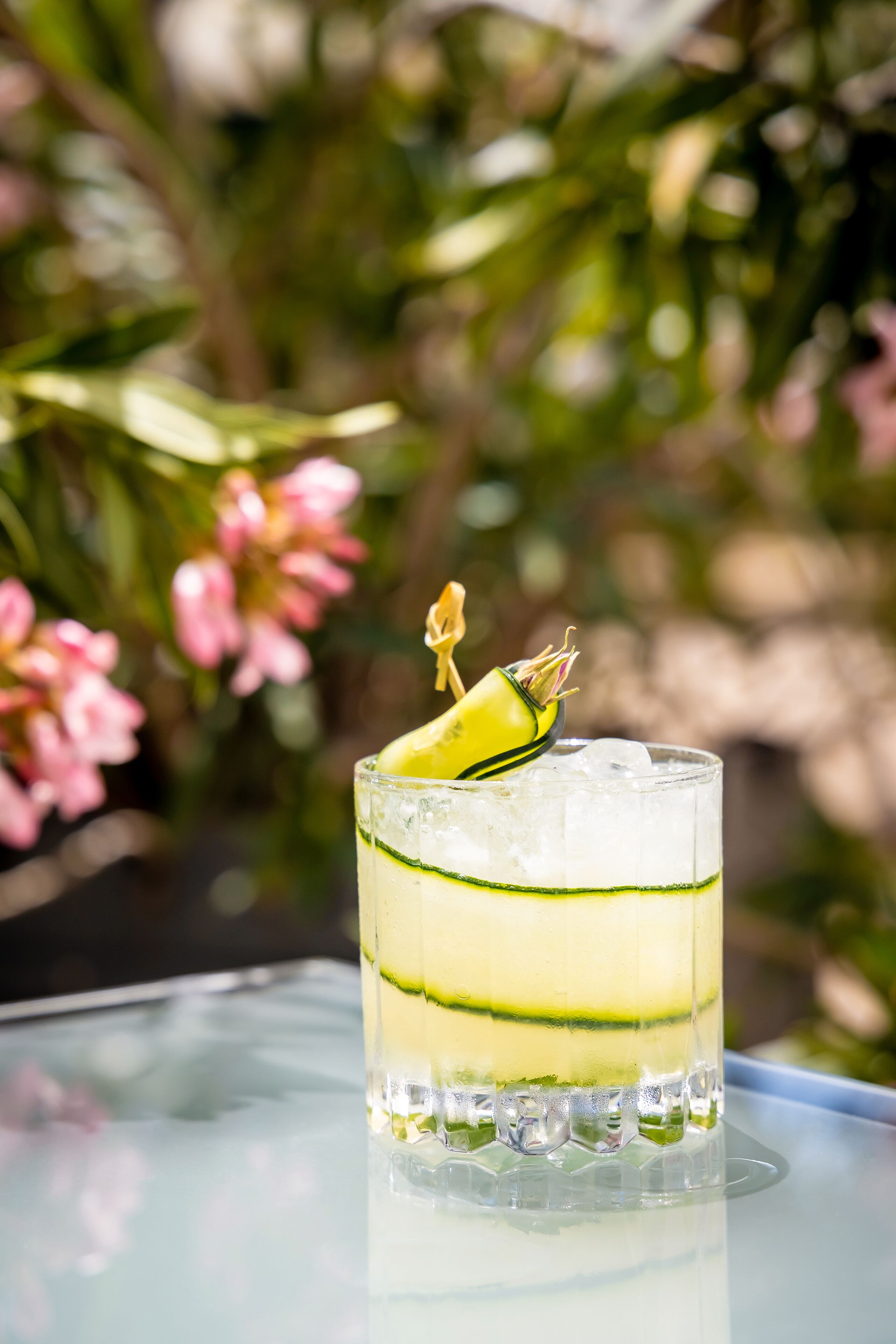 A clear glass with a lime slice garnish on a table with a blurred background of greenery and pink flowers.