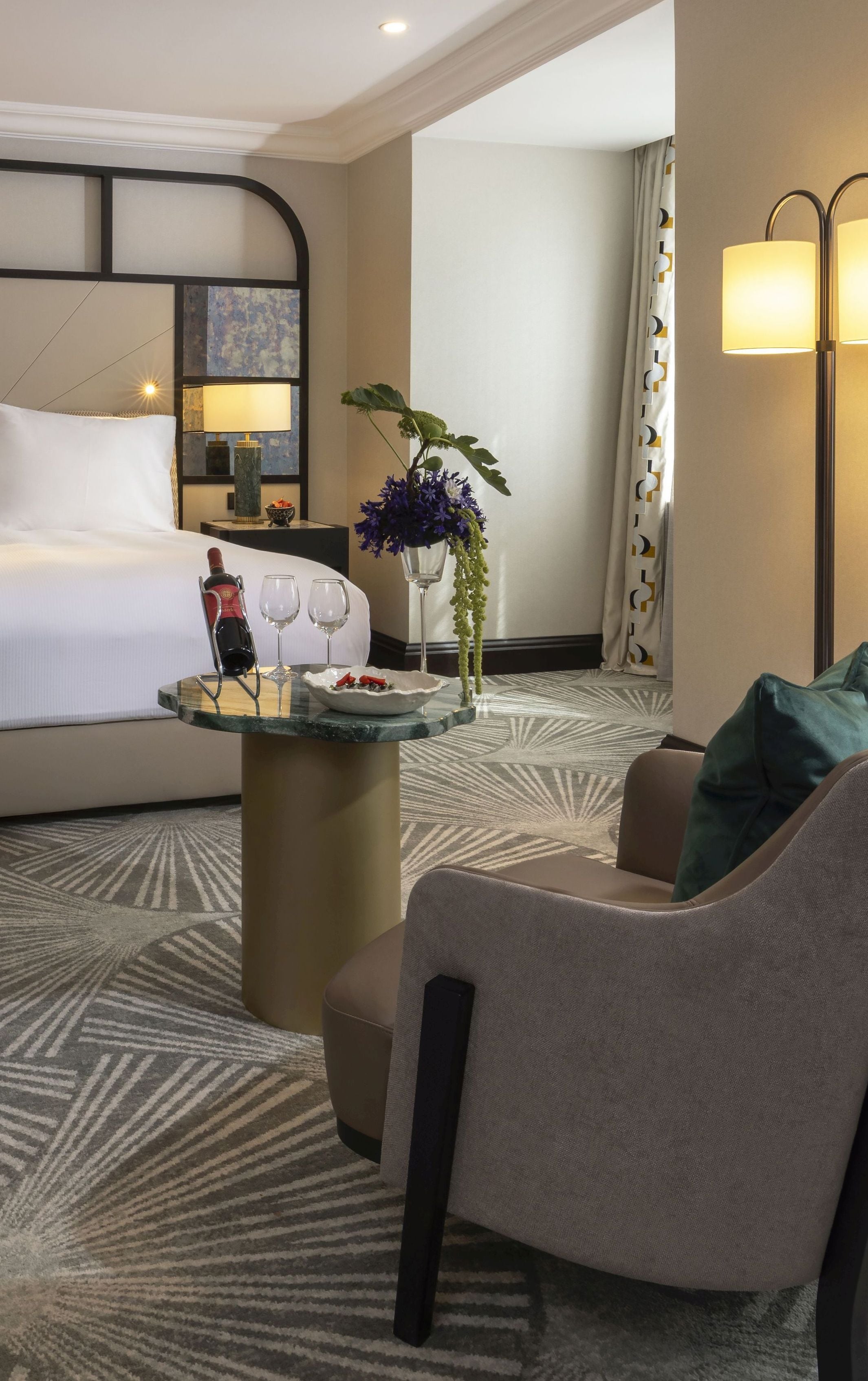 A hotel room with a bed, a seating area with a table, a vase with flowers, and a lamp.