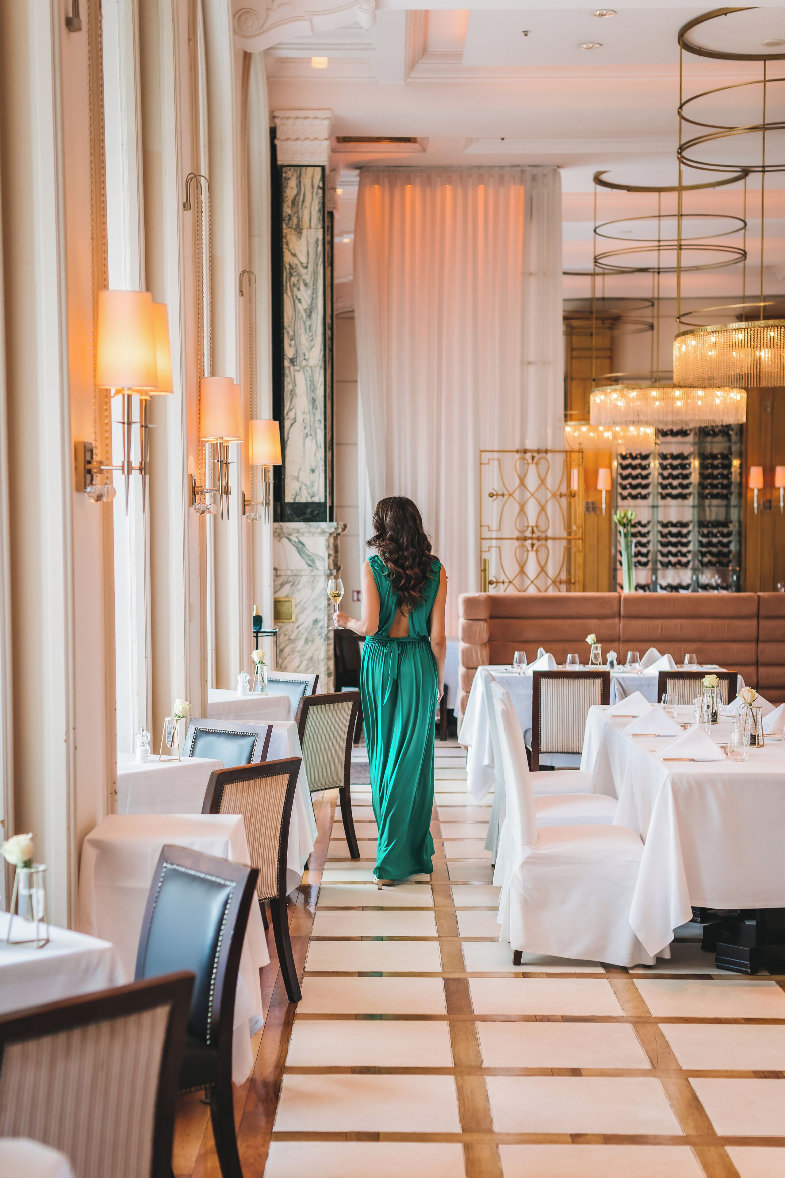 A hotel dining area with tables set for service, a woman in a green dress, and a chandelier.