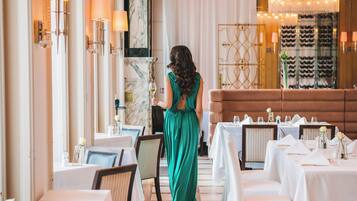 A hotel dining area with tables set for service, a woman in a green dress, and a chandelier.