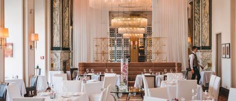 A formal dining room with tables set for service, a central chandelier, and a waiter in the background.