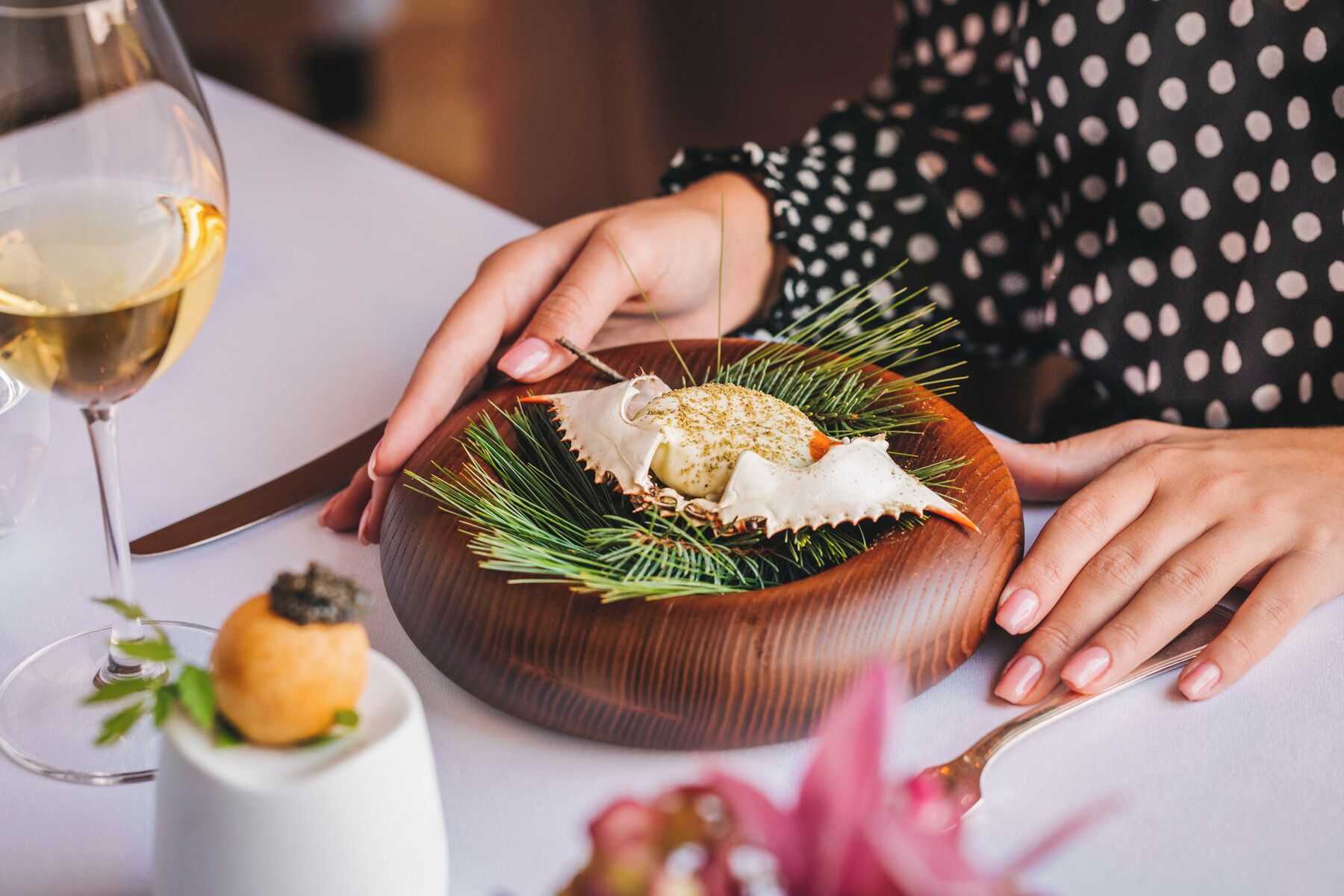 Una persona sostiene un cuenco de madera con un plato de mariscos, una copa de vino y una pequeña taza blanca con una guarnición.